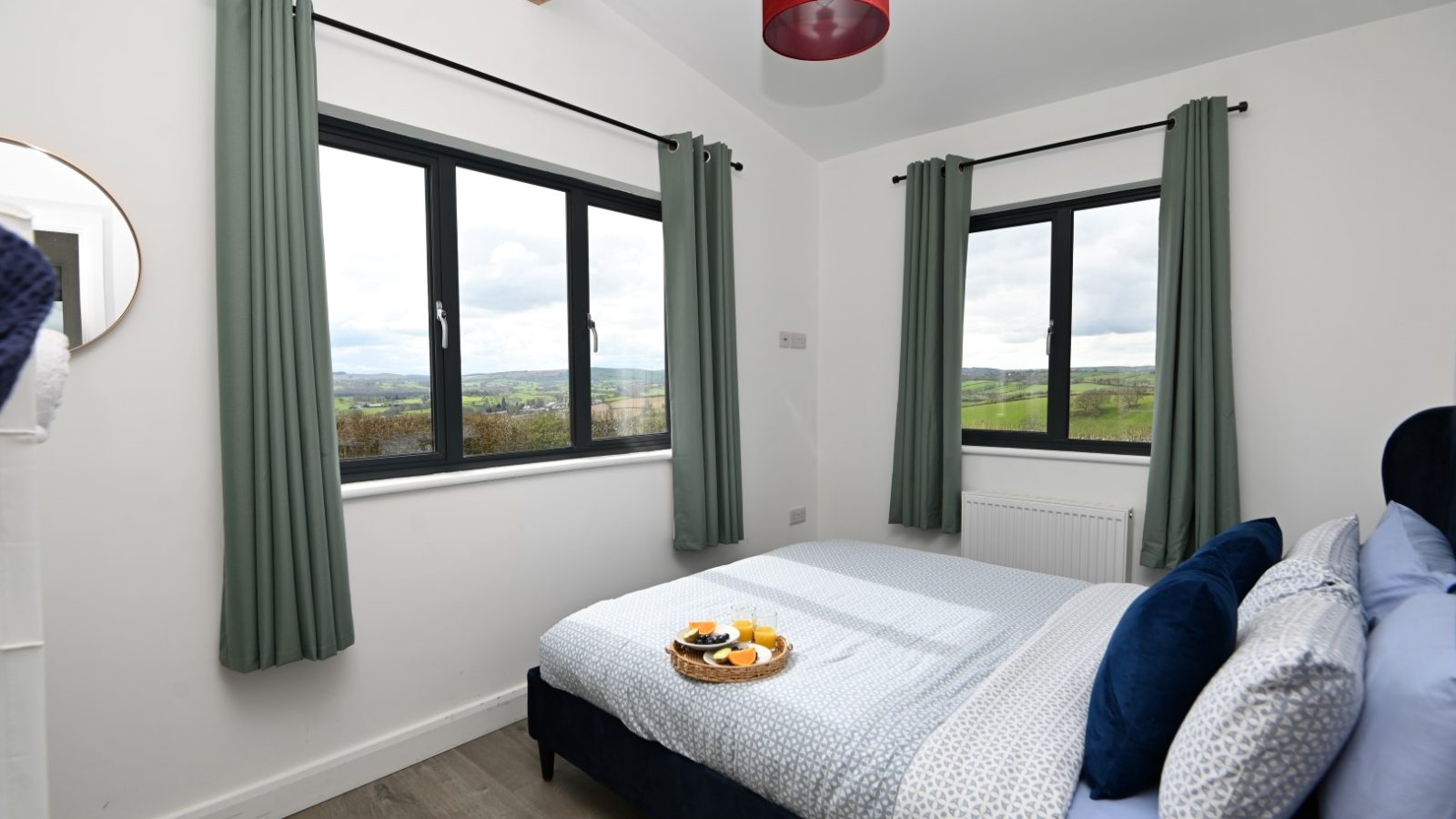 A cozy bedroom at Belan View Lodge boasts large windows revealing a scenic countryside. The room features a neatly made bed with blue and white bedding, a red ceiling lamp, green curtains, and a small tray with breakfast items sitting invitingly on the bed.