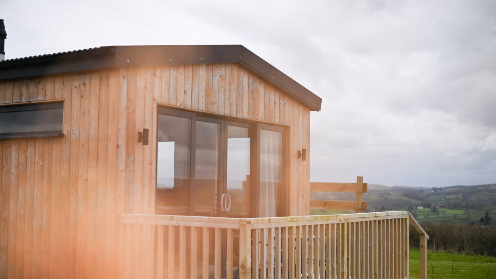 Belan View, a small wooden cabin with large windows, is nestled in a rural landscape. It features a wooden deck with railing. The sky is overcast, and hills are visible in the background. A soft orange tint graces the foreground, adding warmth to this peaceful retreat from The Lodges collection.