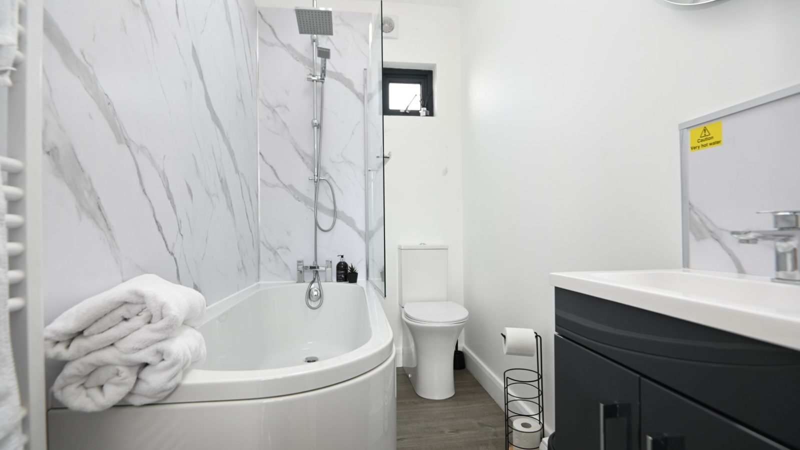 The modern bathroom at Belan View showcases a marble-patterned wall, a sleek bathtub with folded white towels, and a stylish black vanity with a round mirror. The wooden floor and small window add charm while offering natural light, making it the perfect retreat at The Lodges.