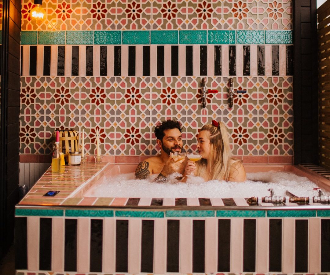 A couple luxuriates in a colorful tiled hot tub, encompassed by an array of bottles. Under the metallic ceiling at Bethnal&Bec, they indulge in drinks, basking in warm lighting that casts a cozy ambiance perfect for unforgettable stays.