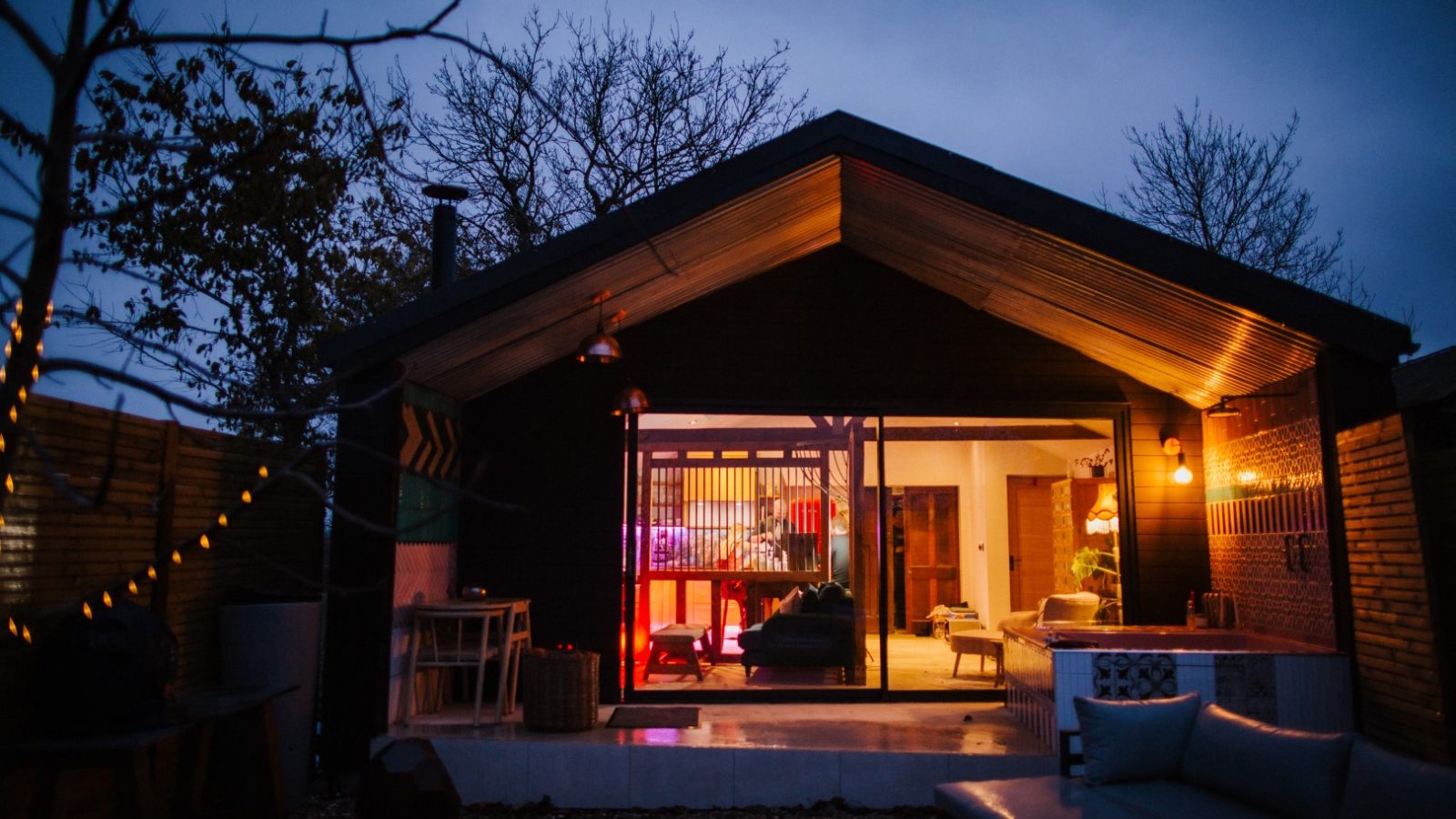 A cozy, modern cabin by Bethnal&Bec boasts large glass windows glowing warmly at dusk. Soft ambient lighting illuminates the interior. Leafless trees and a fence create a tranquil ambiance. Experience luxury stays with a patio and stylish furniture visible in the foreground.