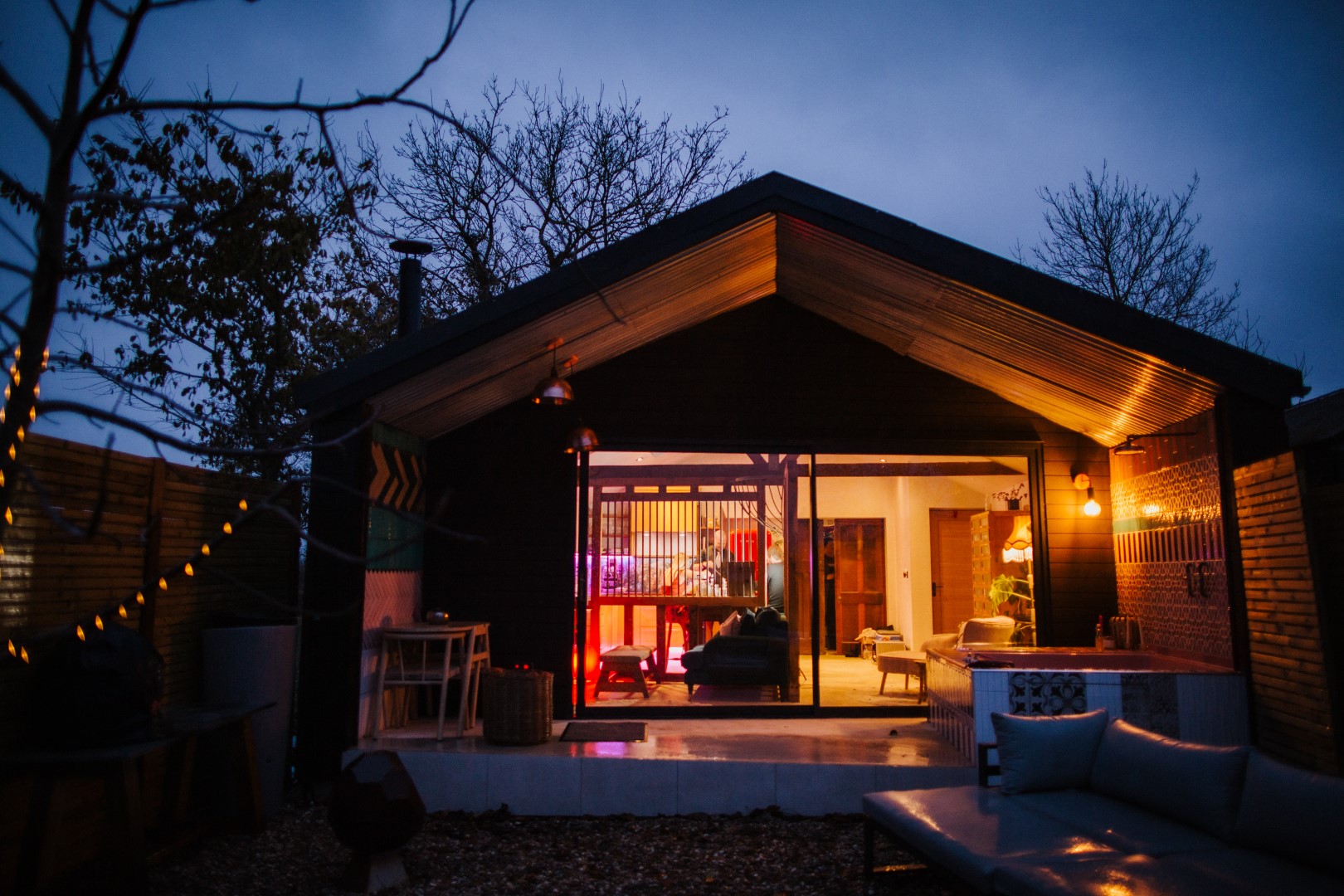 A cozy, modern cabin by Bethnal&Bec boasts large glass windows glowing warmly at dusk. Soft ambient lighting illuminates the interior. Leafless trees and a fence create a tranquil ambiance. Experience luxury stays with a patio and stylish furniture visible in the foreground.