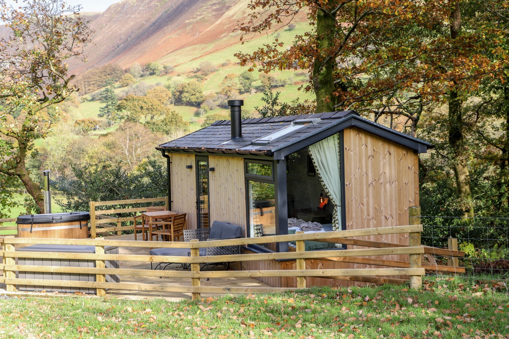 Secluded getaways: A small wooden cabin with a fenced patio, nestled among trees in a hilly, grassy landscape.