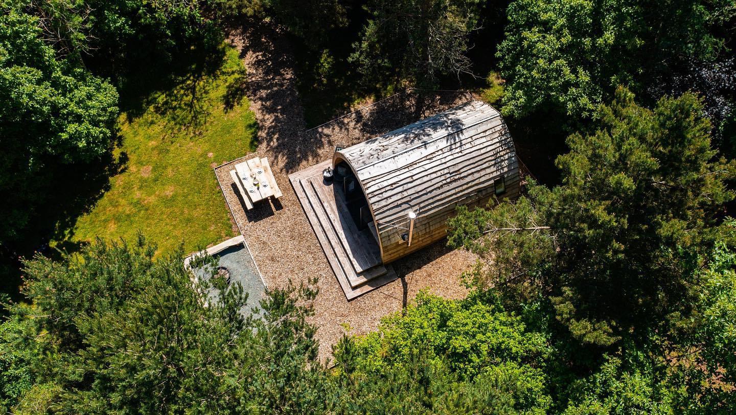 Aerial view of a small, curved wooden cabin nestled in lush woodland. A picnic table sits on a gravel area beside the cabin. Sunlight filters through the surrounding foliage, creating a serene forest setting.