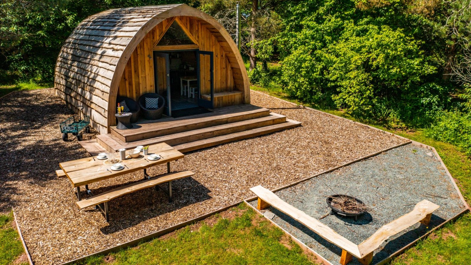 A charming woodland cabin with a curved roof nestles in a forest clearing. In front, there's a picnic table adorned with dishes and a fire pit encircled by log seating. The porch, complete with chairs, is enveloped by the lush greenery of Ffynnonau Annex.