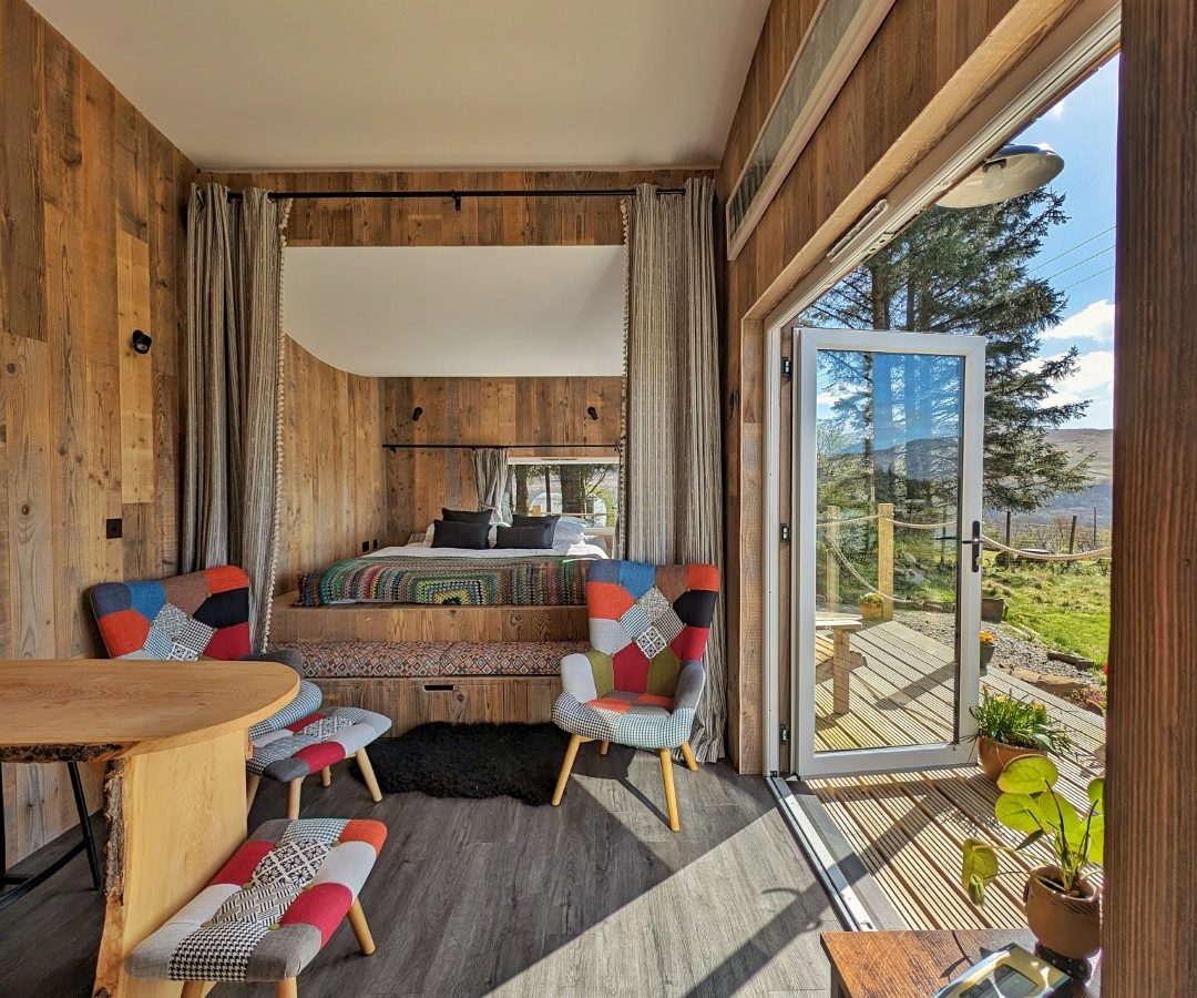 The cozy interior of a Forgotten Coast cabin features a bed with colorful cushions, two matching chairs, and a wooden table. Sunlight streams through the open sliding doors, revealing a deck and scenic views of trees and hills outside.