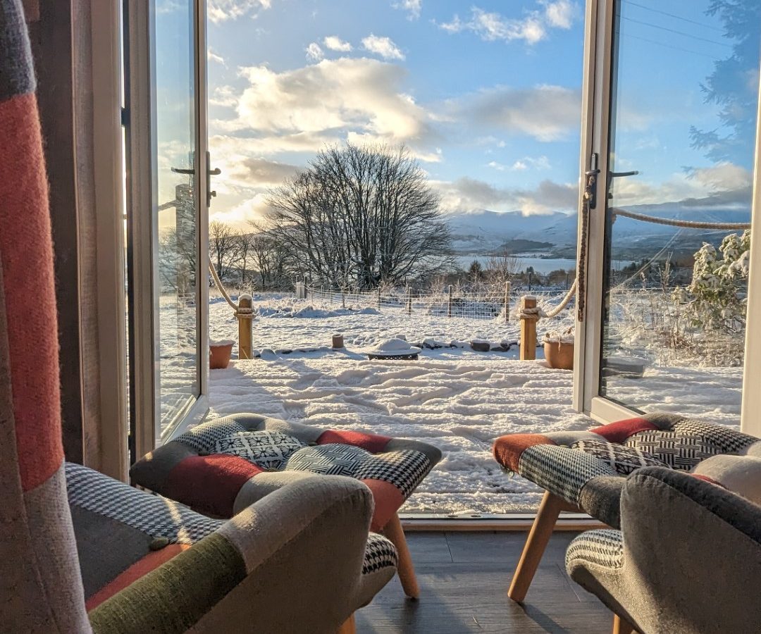 A cozy room with armchairs facing an open door, revealing a snowy landscape outside. A lone tree stands in the center under a bright blue sky, while picturesque mountain cabins dot the backdrop of a majestic range on the iconic Forgotten Coast.