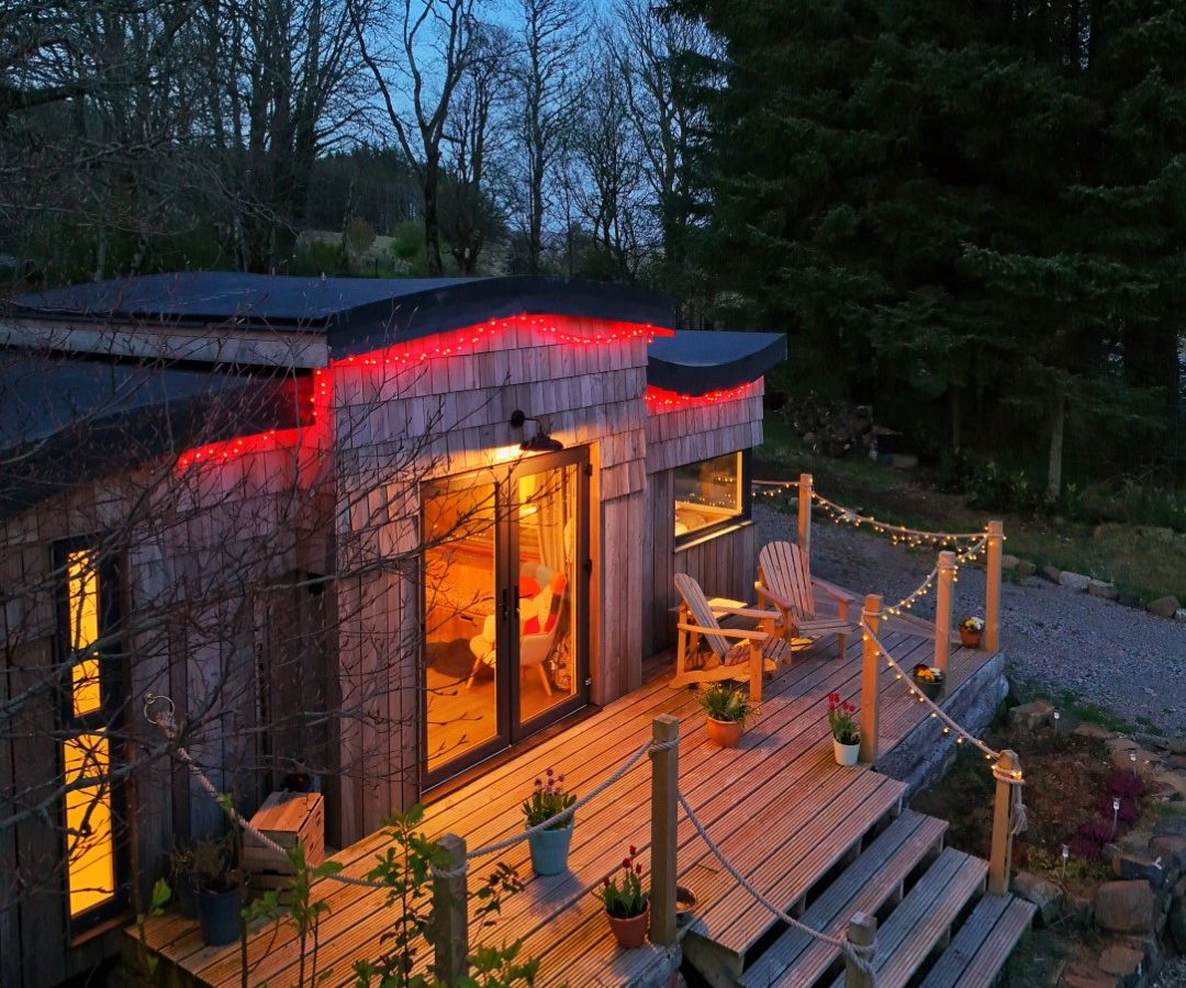 A cozy cabin on the Forgotten Coast features wooden decking warmly lit from within. Red string lights outline the roof, while small lights twinkle along the railing. Adirondack chairs rest on the deck amidst potted plants, with tall trees and a dusky sky providing a serene backdrop.