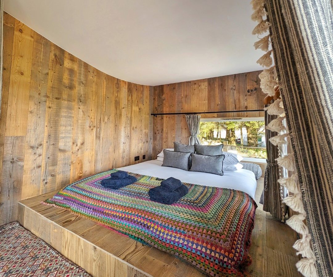 A cozy bed on a wooden platform with a colorful, patterned blanket and two folded towels, this room feels like the perfect retreat in one of those charming Forgotten Coast cabins. Wooden walls and patterned curtains frame a window view of tree-lined tranquility.