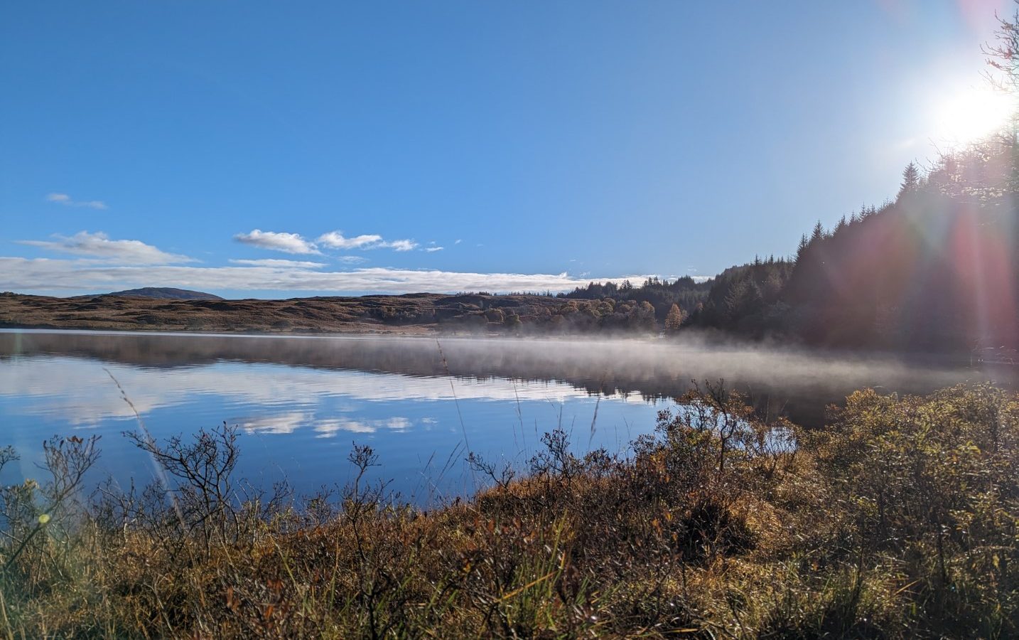 A serene lake scene with a clear blue sky and bright sun. Mist rises from the water's surface, where quaint cabins dot the shore. Grassy terrain and distant hills frame the tranquil landscape, evoking memories of a peaceful retreat along the Forgotten Coast.