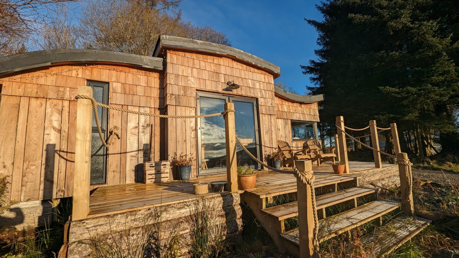 A modern wooden cabin with large glass doors stands in a wooded area on the serene Forgotten Coast. It has a spacious deck with potted plants and a rope railing. The sky is clear and blue, reflecting on the cabin's windows, enhancing its peaceful coastal charm.