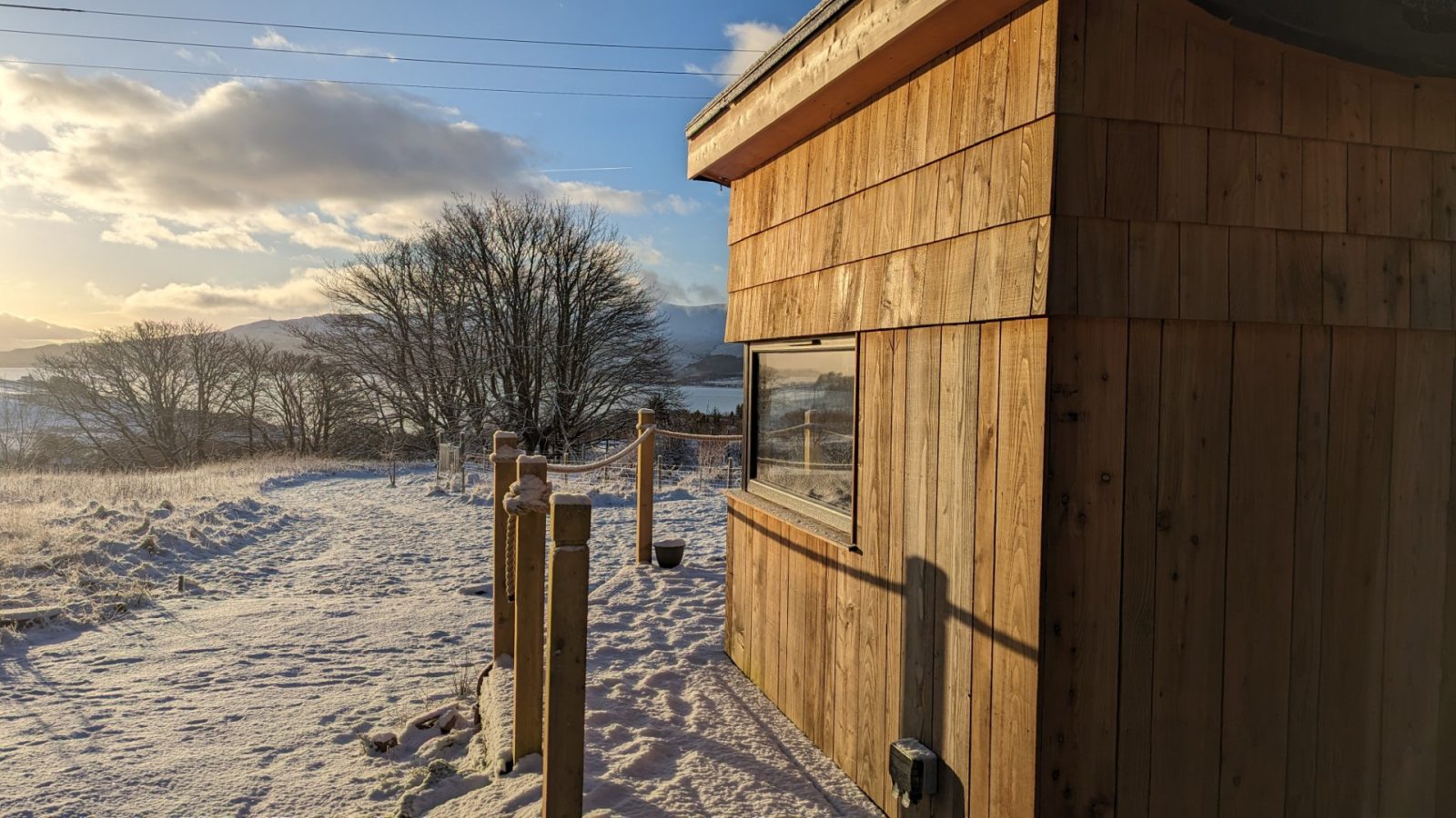 A small wooden cabin rests in a snowy landscape on the edge of the Forgotten Coast, with a path leading away. A tree stands nearby, and mountains are visible in the background under a partly cloudy sky. Sunlight casts warm tones on the tranquil scene.