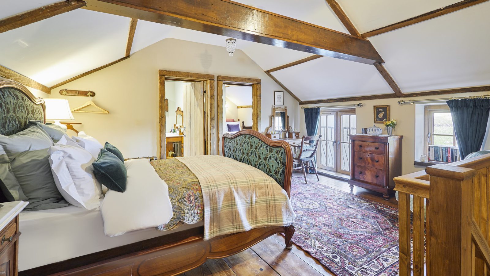 A cozy bedroom in Black Mountain with a vintage wooden bed and a colorful rug on wooden floors. Exposed beams add rustic charm. A small desk and window are in the corner, offering natural light. The room features warm tones and classic furniture, perfect for serene escapes.