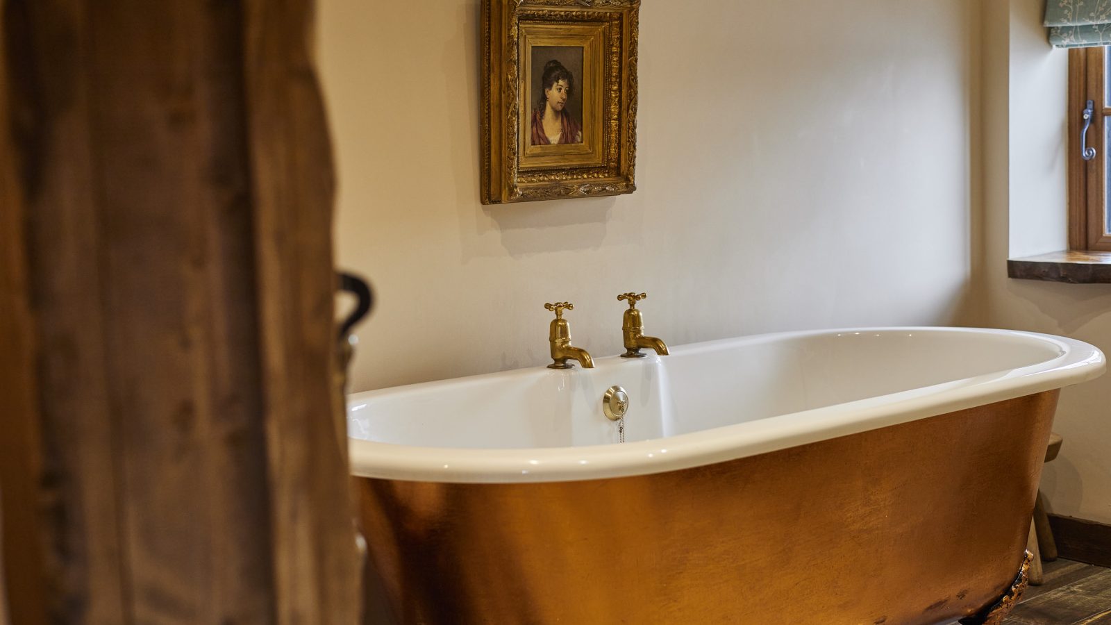 A bathroom at Gablebach features a vintage-style freestanding copper bathtub with brass taps. Above the tub, a framed portrait graces the wall. A wooden beam and a window with a short curtain are partially visible, reminiscent of the charm found at Black Mountain Escapes.