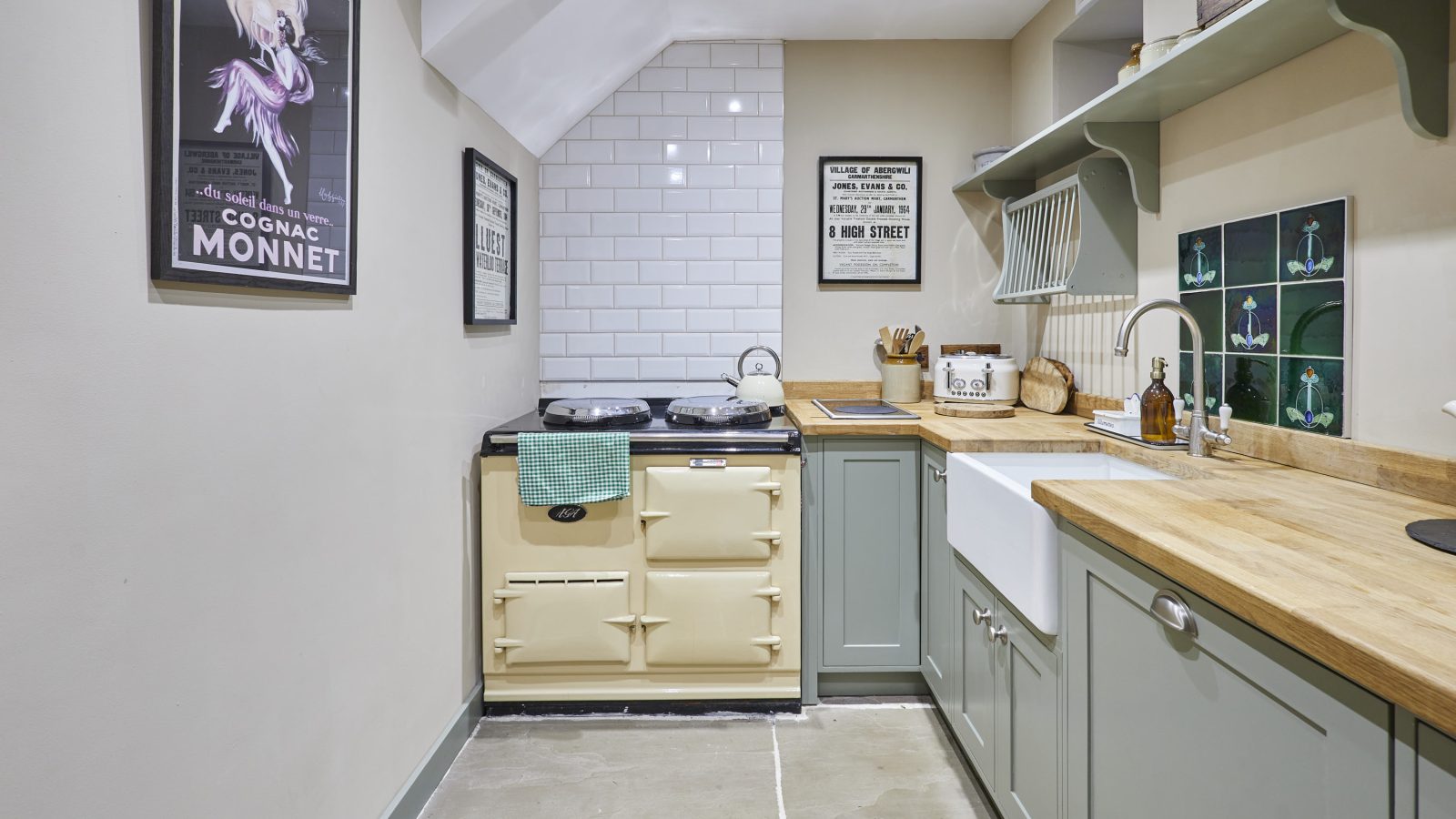 A cozy kitchen with beige and green cabinets, a farmhouse sink, and a retro-style stove evokes a Black Mountain retreat. The walls feature white subway tiles and decorative posters. Wooden countertops and shelves hold dishes and kitchenware, while stone flooring completes the rustic look.