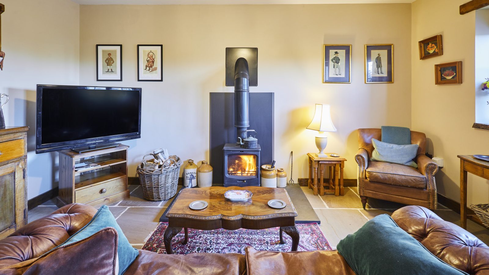 Cozy living room with a leather sofa, armchair, and wooden coffee table. A wood-burning stove from Black Mountain Escapes is lit against the back wall. The room features framed artwork, a television, and a woven basket with firewood. A soft lamp illuminates the corner.