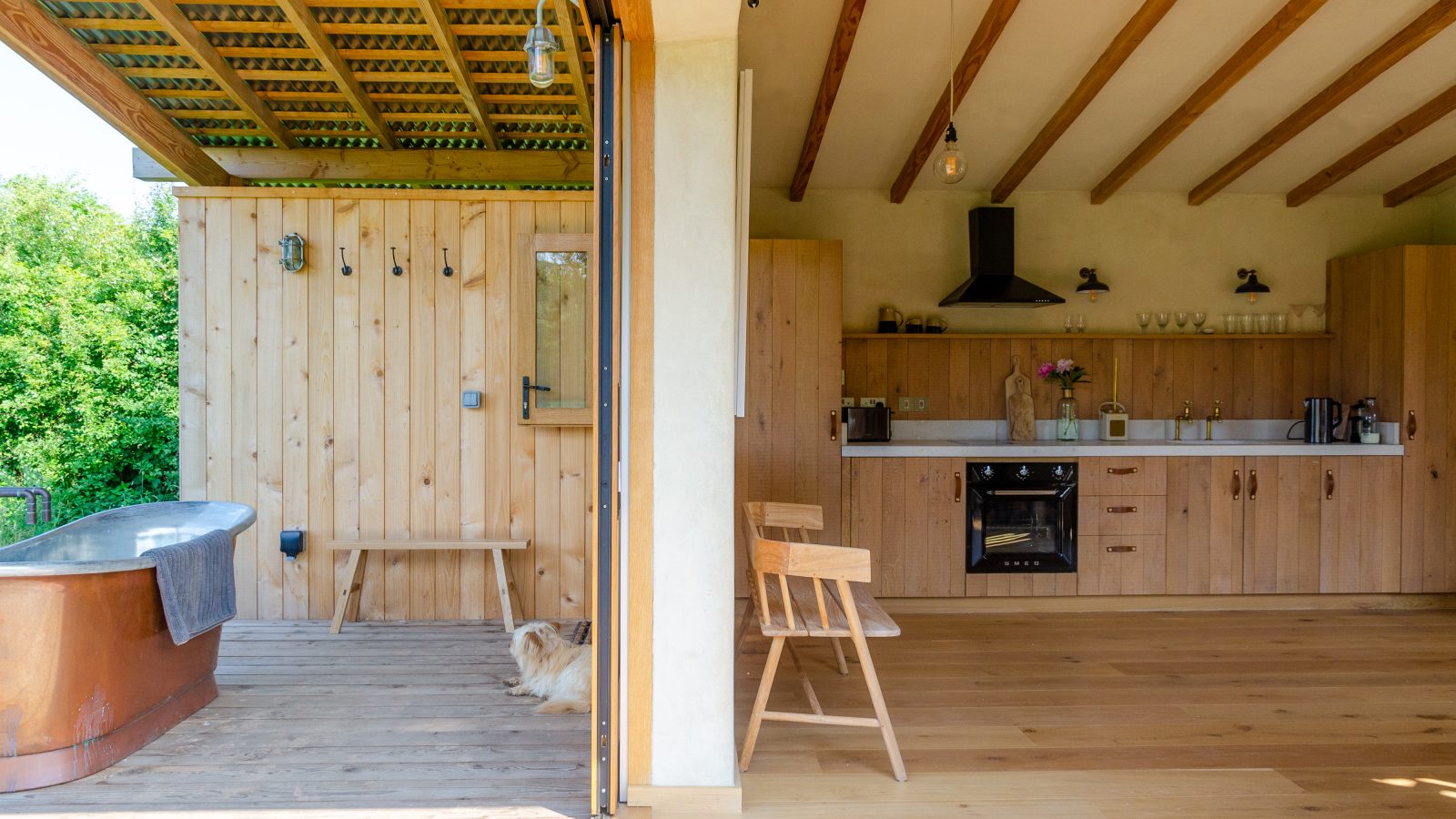 The Holly Water Cabin offers a cozy open-air space with light wooden cabinets in the kitchen, an indoor plant, and a dog resting beside a wooden bathtub on the deck. Exposed beams grace the roof while the deck overlooks lush greenery.