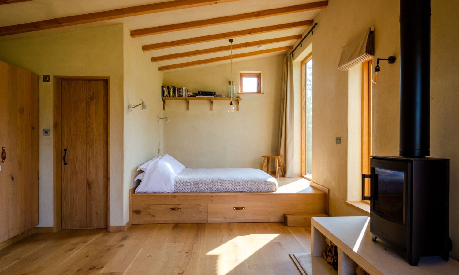 The Holly Water Cabin features a cozy room with a bed nestled into a wooden alcove on the left, and natural light streams through the window. A black wood stove graces the right side, with exposed wooden beams overhead and beige walls harmonizing with the wooden floor.