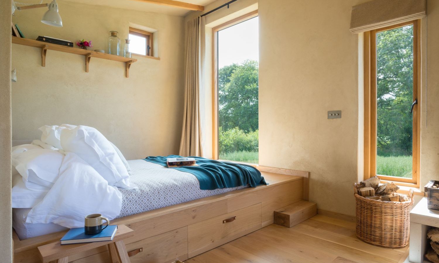 A cozy bedroom in the Holly Water Cabin features a wooden bed adorned with white linens and a teal blanket. Large windows offer views of lush greenery. A wicker basket holds firewood beside the bed, while a small wooden stool with a mug sits nearby, adding to the rustic charm.