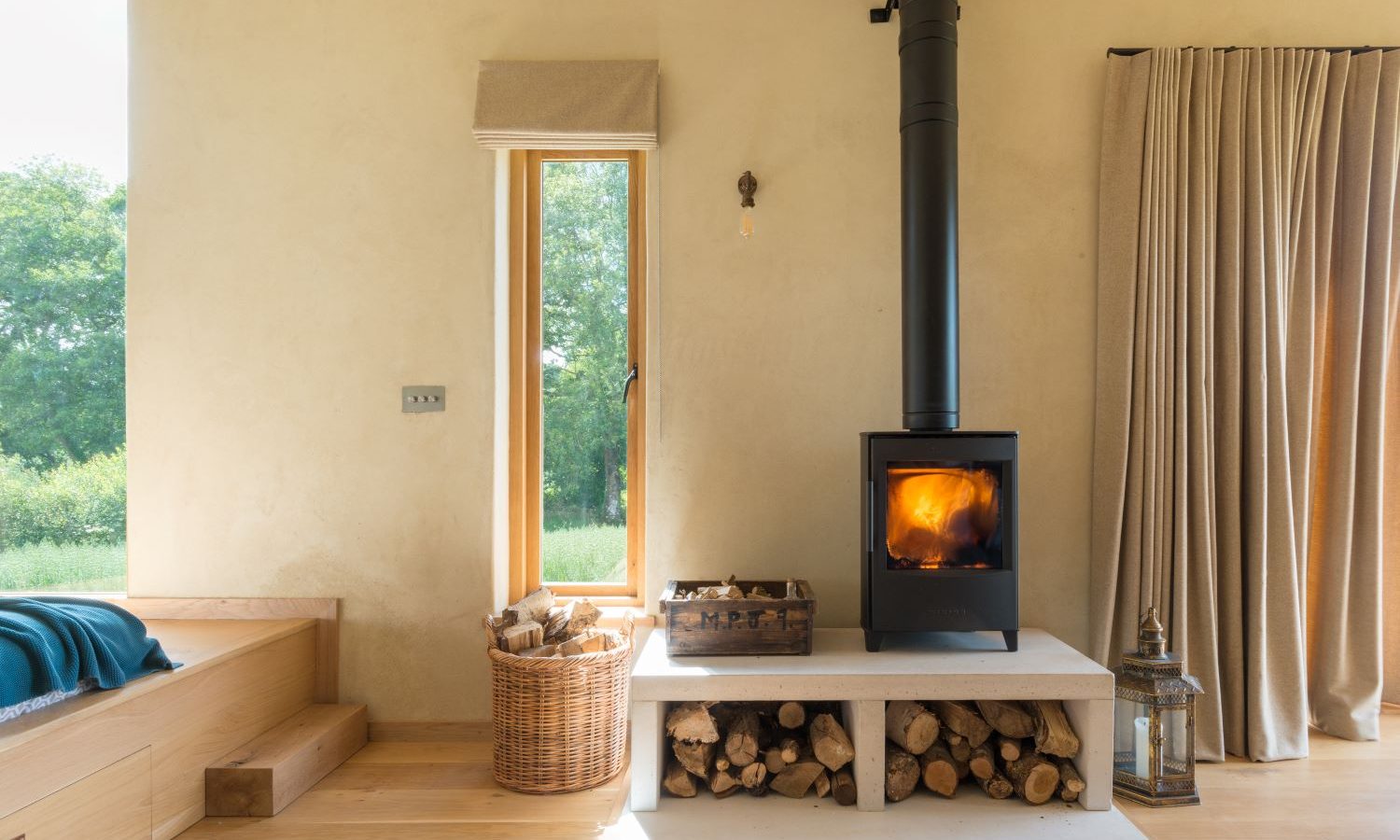 A cozy room in the Holly Water Cabin features a modern black wood stove emitting a warm glow. The stove sits on a white platform with firewood stored underneath. A wicker basket with logs rests beside a tall window with beige curtains, letting natural light filter in.