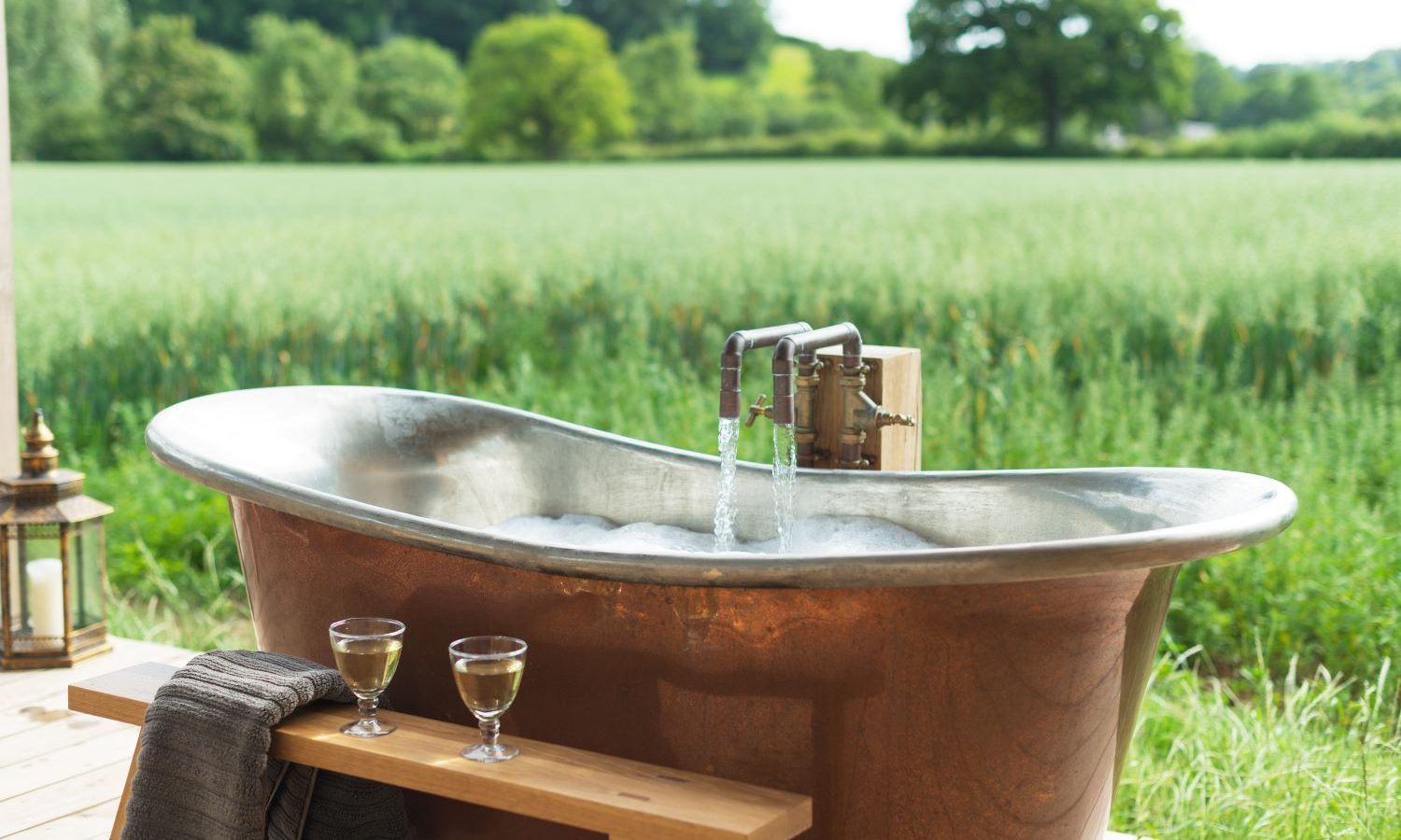 A luxurious copper bathtub with water running into it graces the deck of Holly Water Cabin, overlooking a lush green field. Two glasses of white wine and a towel sit on a wooden ledge beside the tub, while trees and a clear sky create a serene backdrop.