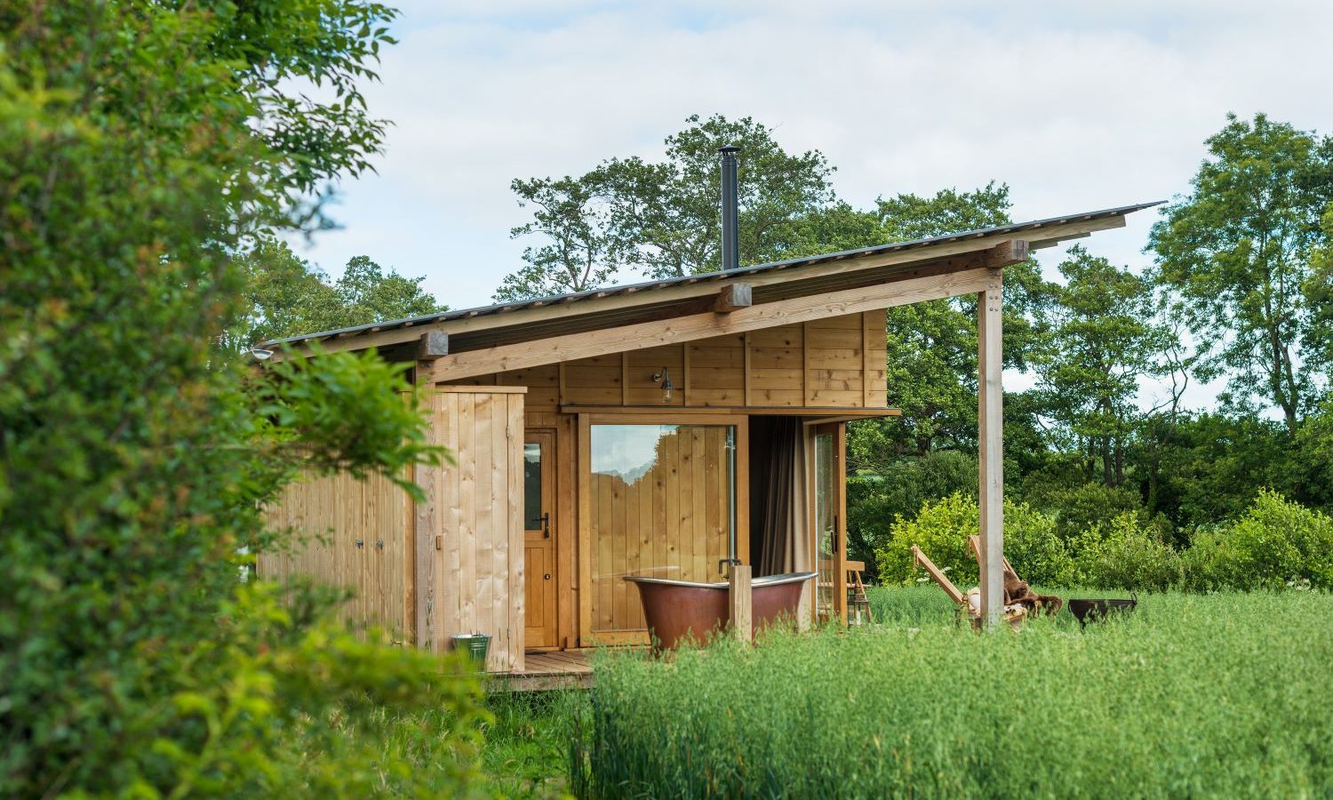 Holly Water Cabin, a small wooden retreat with a sloped roof, is nestled among tall grass and trees. A red bathtub graces the porch alongside a wooden chair. The cloudy sky adds to the serene and peaceful atmosphere of this enchanting hideaway.