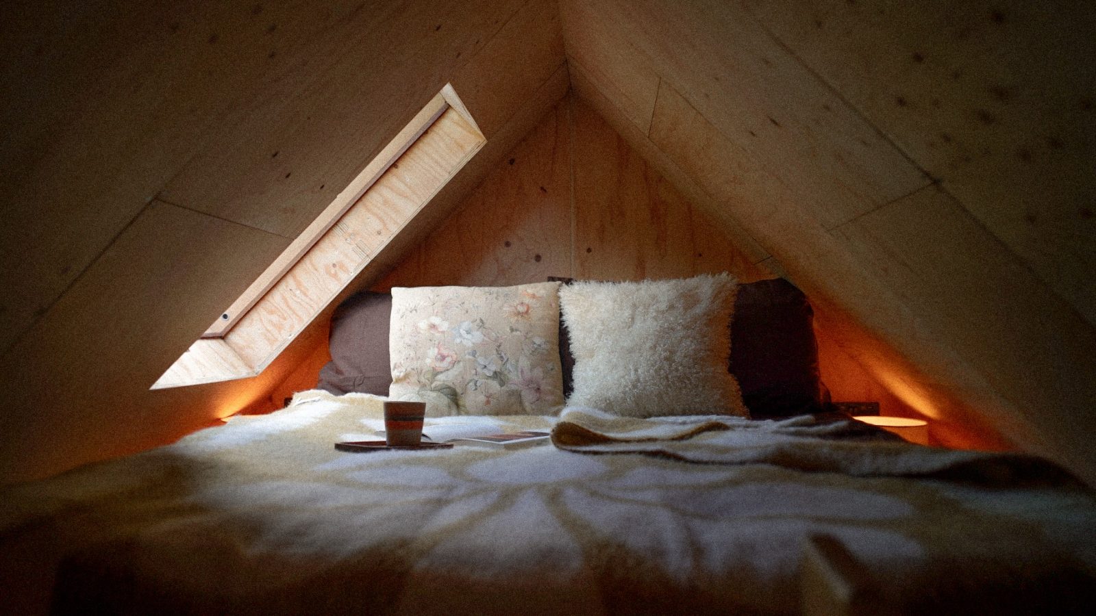 A cozy attic bedroom designed by Korkhouse features a sloped wooden ceiling and a skylight. The bed, adorned with soft pillows and a plush blanket, invites relaxation. A small cup and notebook rest on the bed, bathed in warm ambient lighting for a truly tranquil escape.