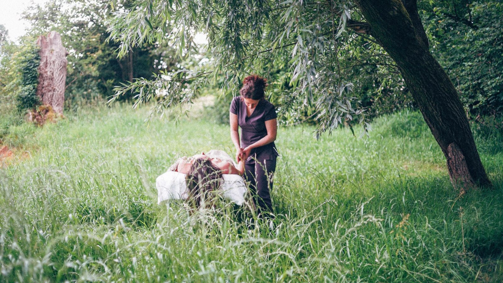 A person from Korkhouse kneels beside another lying on a blanket in a grassy field. They are enveloped by trees and greenery, crafting a peaceful, natural setting.