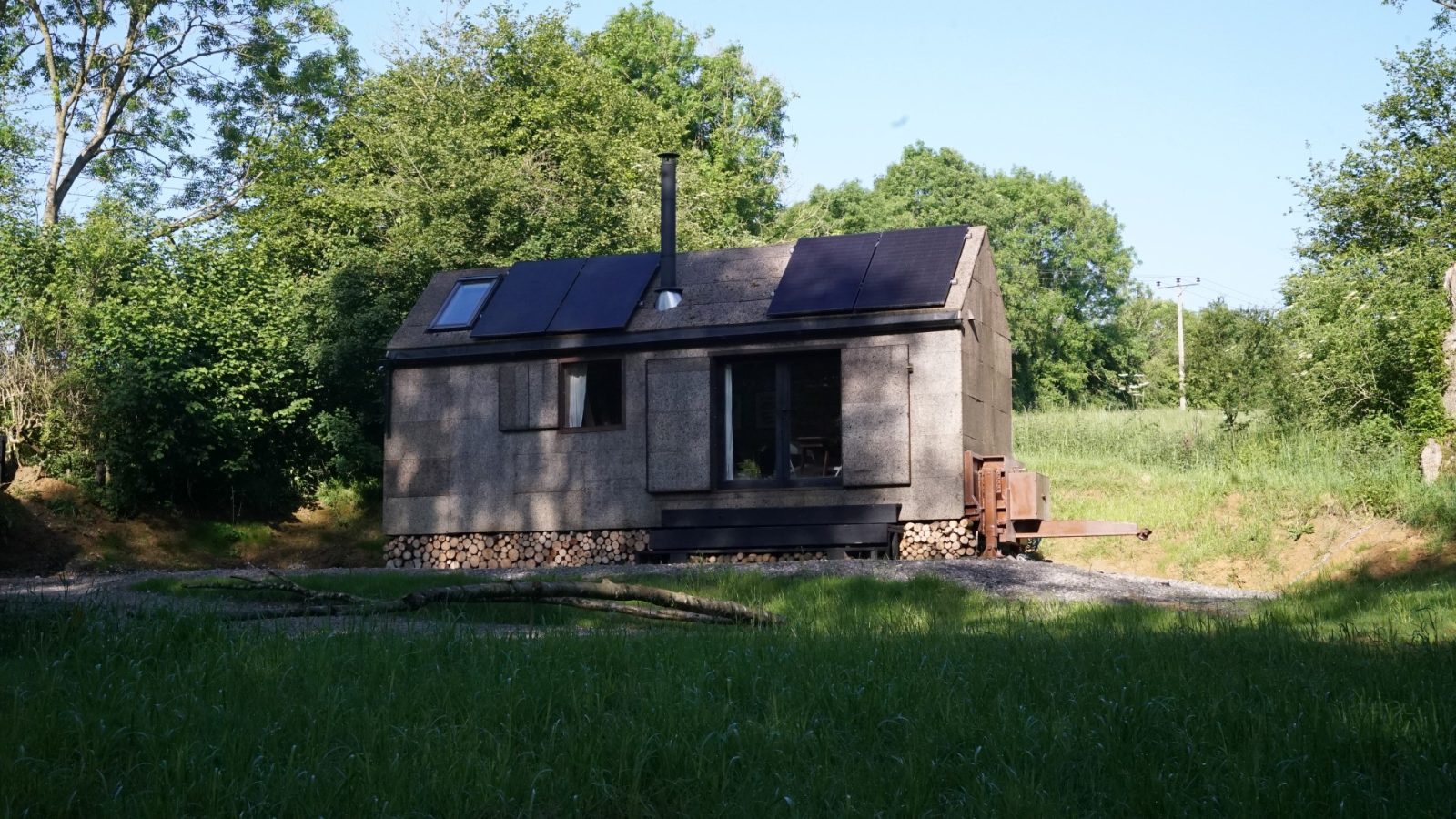 A small, rustic Korkhouse cabin with solar panels is situated in a grassy clearing surrounded by trees. The cabin features a sloped roof, a chimney, and wood storage underneath. The scene is bathed in natural sunlight.