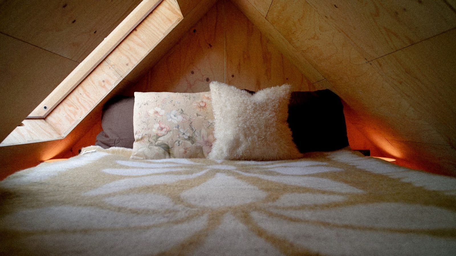 A cozy bed in a wooden loft with slanted walls, nestled within the innovative Korkhouse design. The bed features floral and fluffy pillows, and a patterned blanket. Warm lighting creates a snug atmosphere, while a skylight above partially illuminates the space.