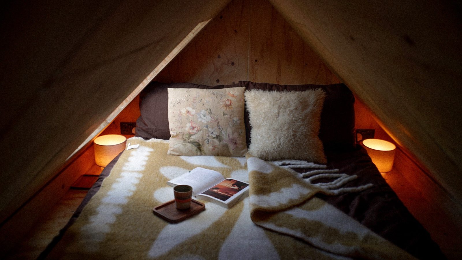 A cozy attic bedroom designed by Korkhouse features soft lighting and a neatly made bed adorned with floral and fluffy pillows. A tray with a mug and open book sits on a patterned blanket, while two small lamps provide the space with a warm, inviting glow.