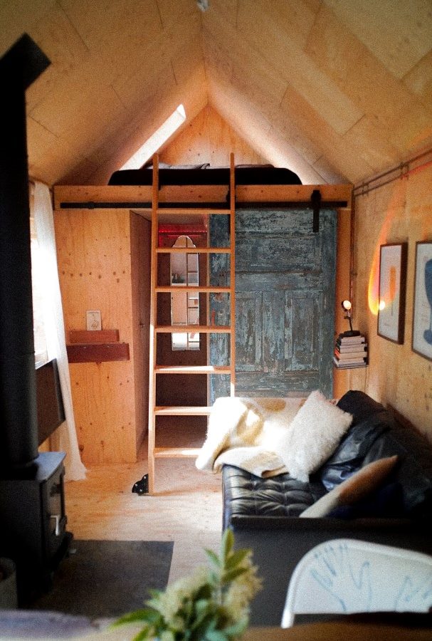 The cozy interior of this Korkhouse tiny home features a rustic loft with a wooden ladder, a black leather couch adorned with pillows and a blanket, and a small wood stove. Framed art graces the walls, while soft lighting creates an inviting warmth throughout.