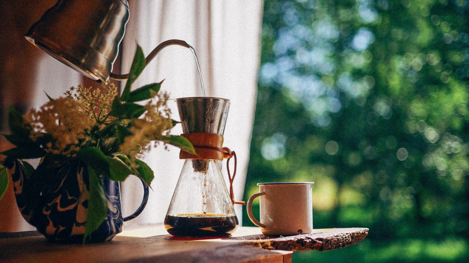 A Korkhouse kettle elegantly pours water into a Chemex coffee maker adorned with a wooden collar on a wooden table. Nearby, a ceramic mug rests beside a floral teapot as sunlight filters through the window, casting gentle rays on the blurred greenery outside.