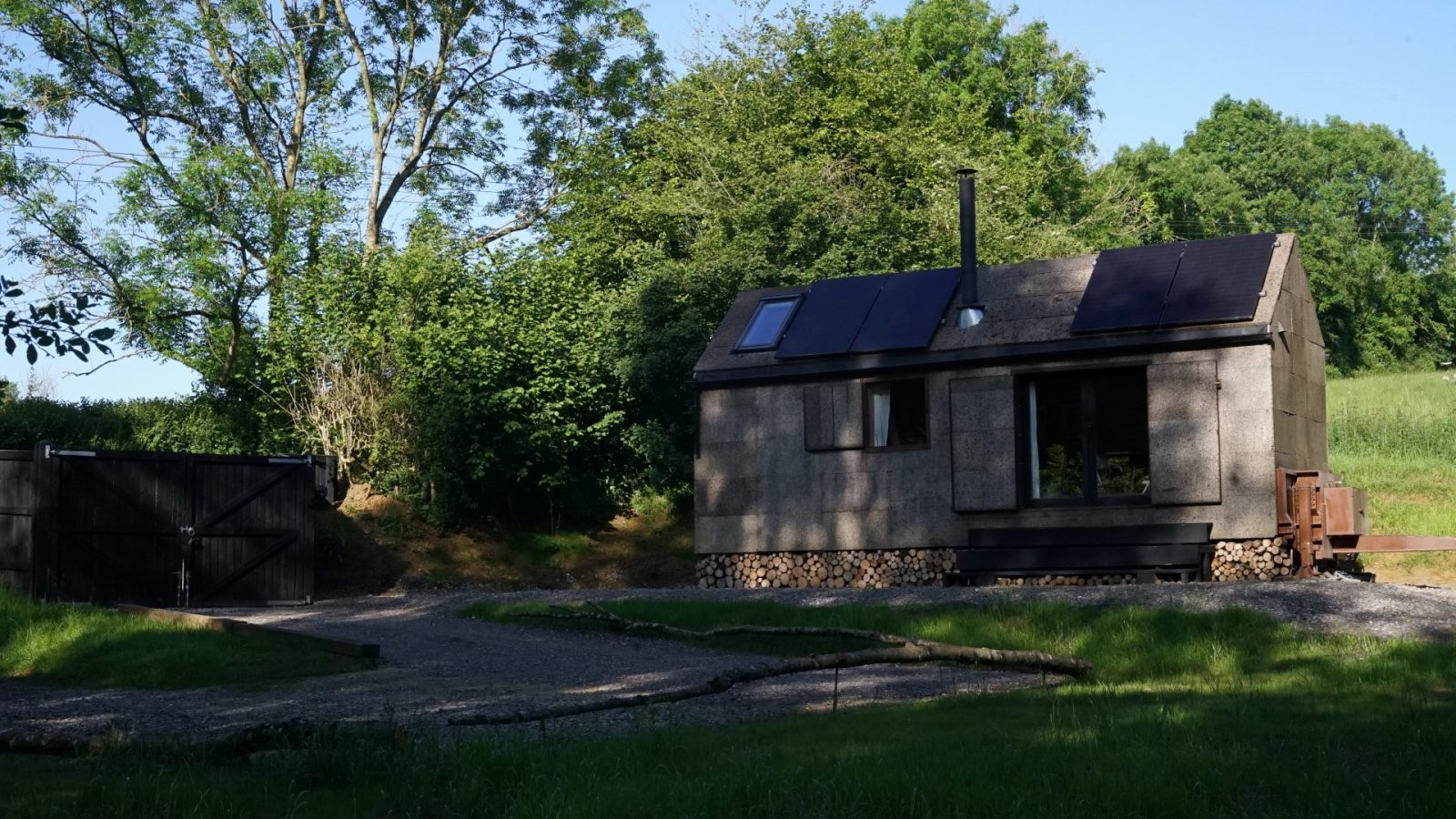 A small, modern cabin with solar panels, designed by Korkhouse, sits in a lush green setting. Surrounded by trees and featuring large windows and a rustic design, it has a path leading to its entrance. A storage shed is visible to the left.