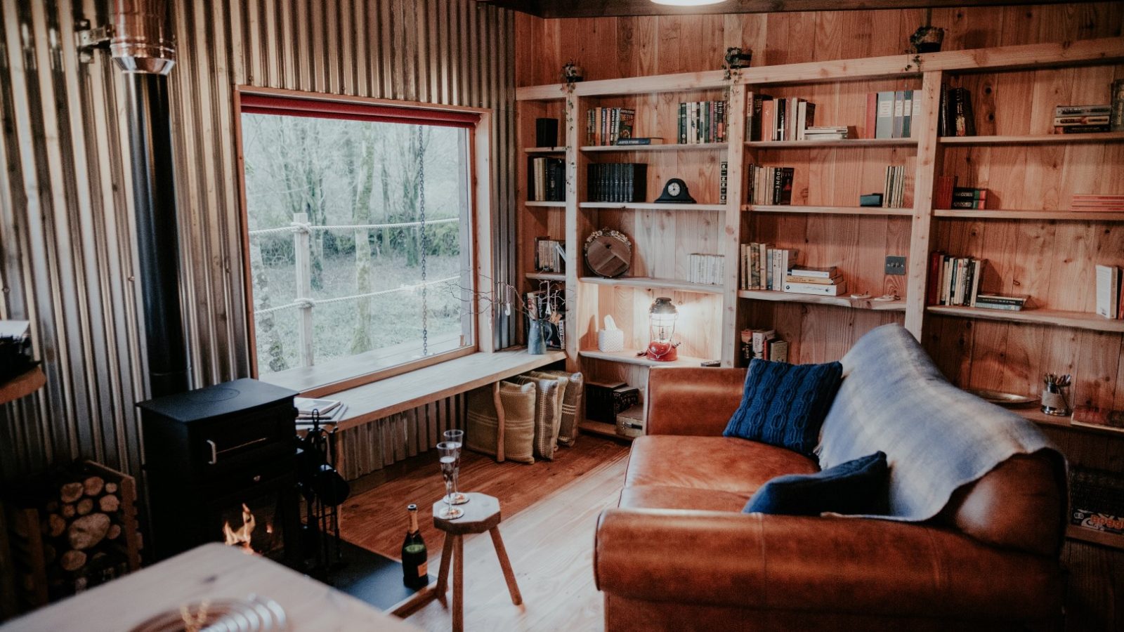 The cozy wooden cabin interior at Little Menherion features a brown leather sofa adorned with blue cushions and a blanket. Bookshelves line the walls, and a window overlooks the forest. A wood stove warms the corner, while various items rest on a small round table.