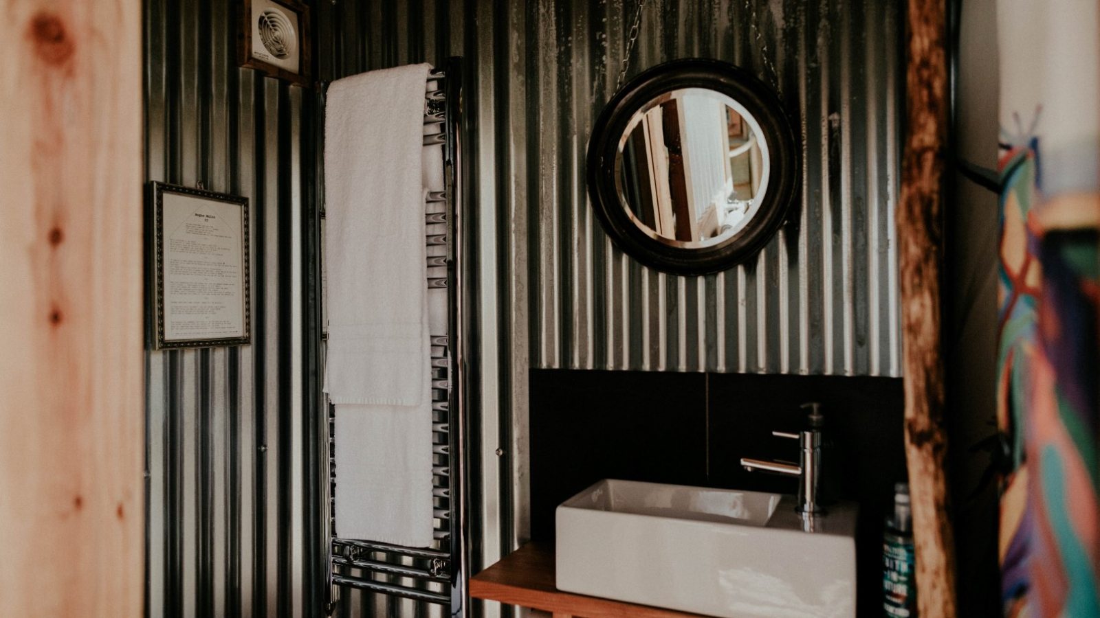 In the quaint Little Menherion bathroom, corrugated metal walls frame a round mirror and a white rectangular sink with a black faucet on a wooden counter. A towel hangs on a wall-mounted rack, while framed text adds charm to the setting.