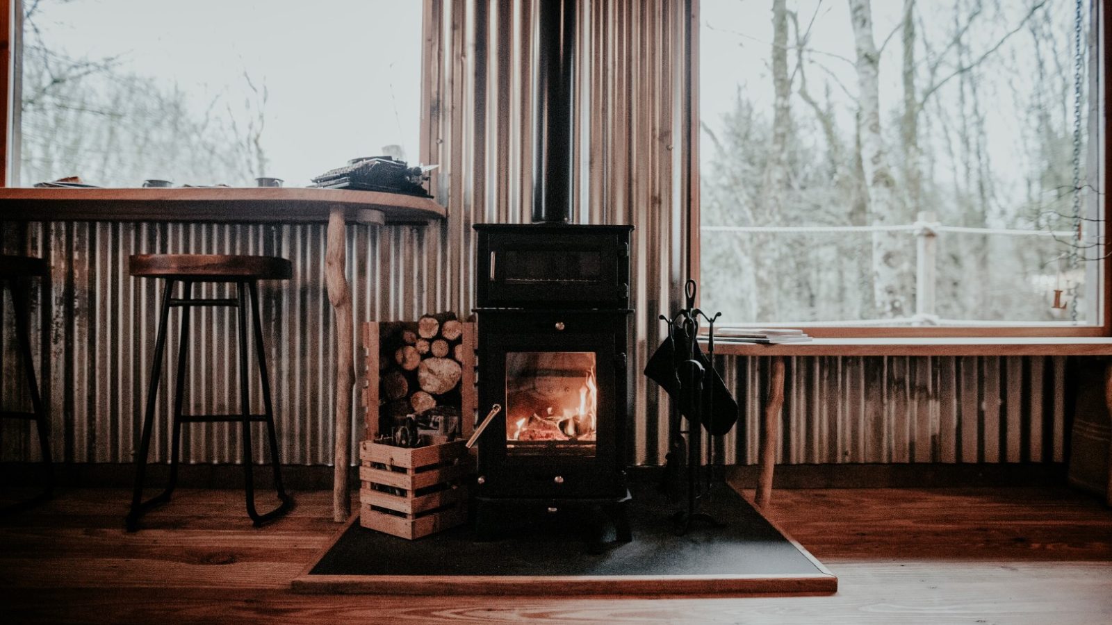 A cozy room in Little Menherion features a small black wood stove burning amidst log supplies. Wooden stools stand by a narrow table with large windows framing the bare trees outside. Inside, the wood flooring and corrugated metal walls create a rustic charm.