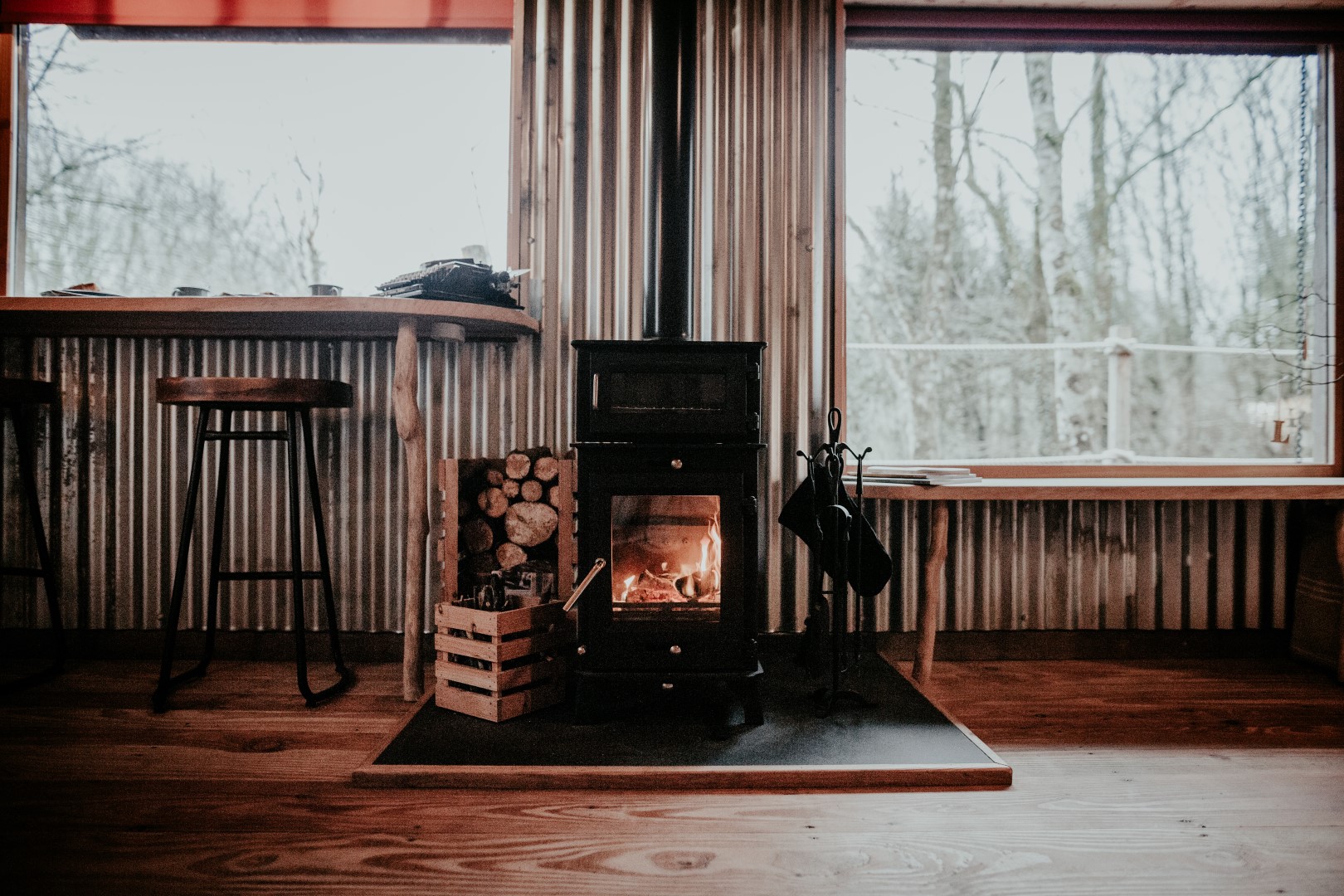 A cozy room in Little Menherion features a small black wood stove burning amidst log supplies. Wooden stools stand by a narrow table with large windows framing the bare trees outside. Inside, the wood flooring and corrugated metal walls create a rustic charm.