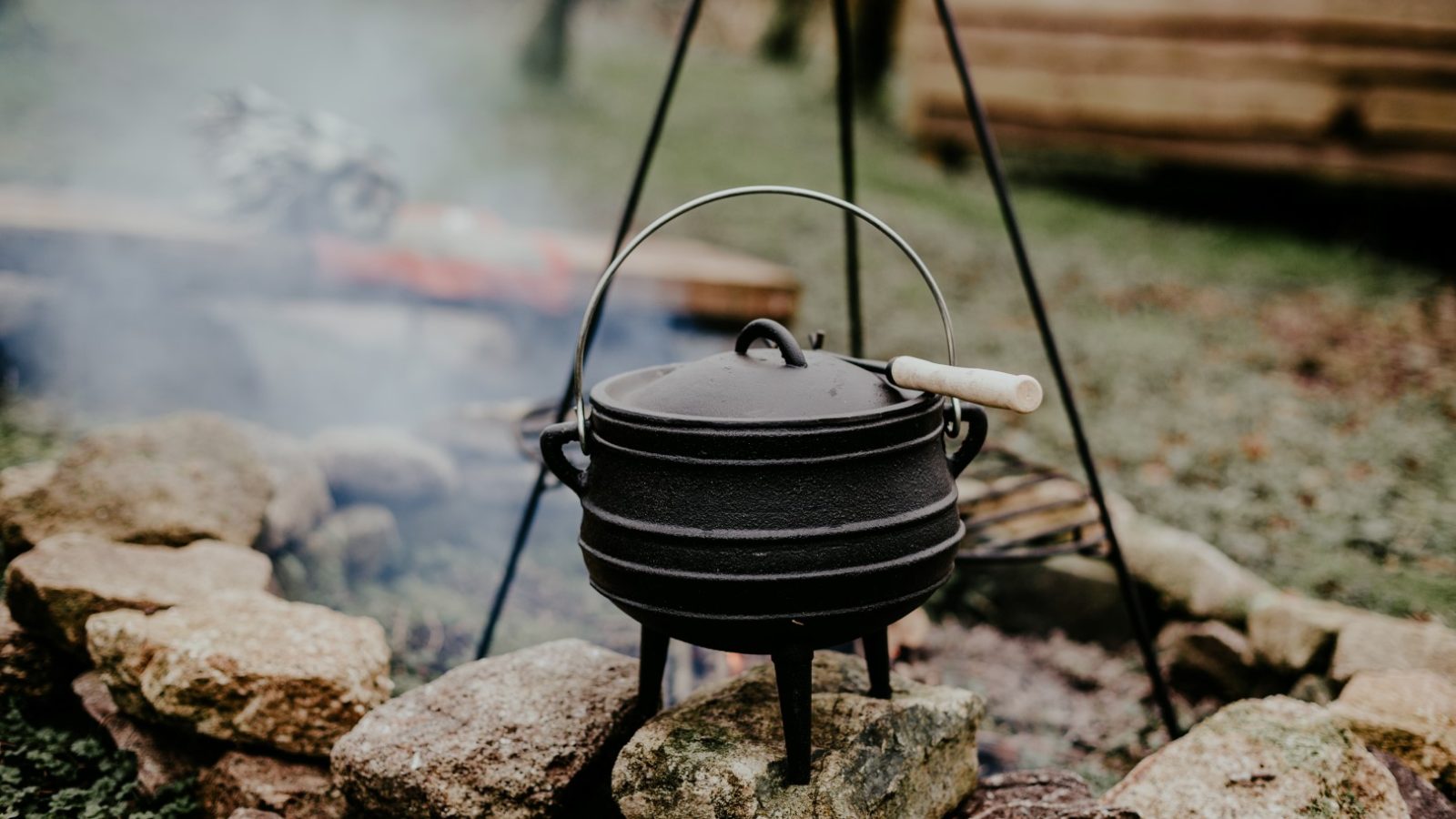 A cast iron pot hangs over an open campfire near Little Menherion, surrounded by a circle of rocks. Smoke gently rises from the fire, and a wooden structure is visible in the background. The scene exudes a rustic, outdoor charm unique to this quaint location.