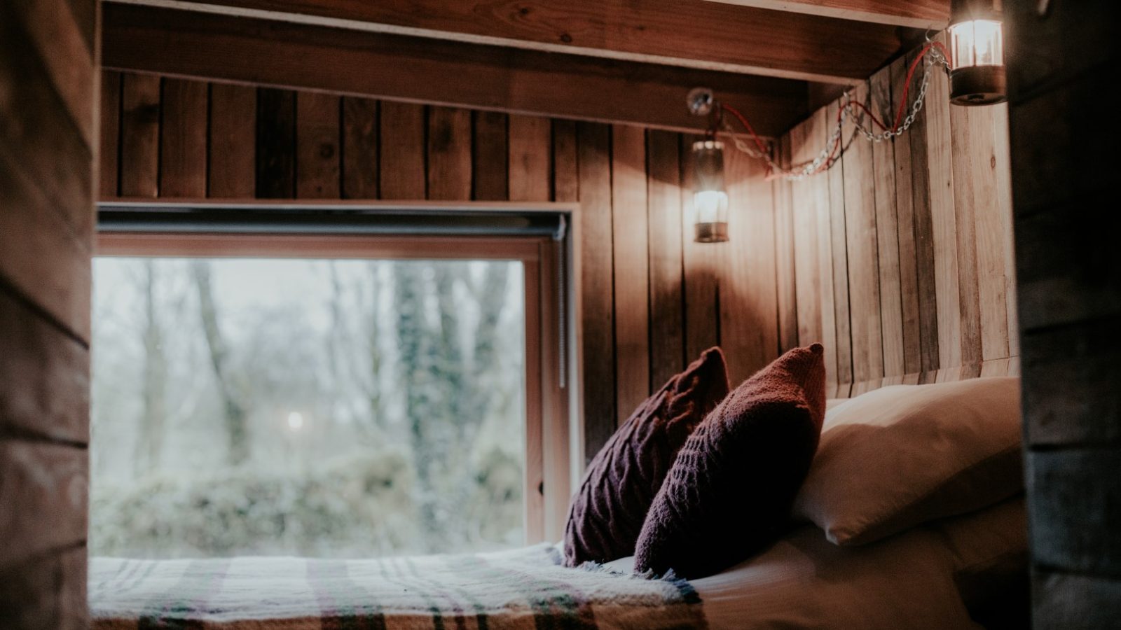 Cozy wooden bedroom at Little Menherion features a large window and soft lighting. A bed with pillows and a blanket is nestled under exposed wooden beams, creating a warm, inviting atmosphere with views of the trees outside.