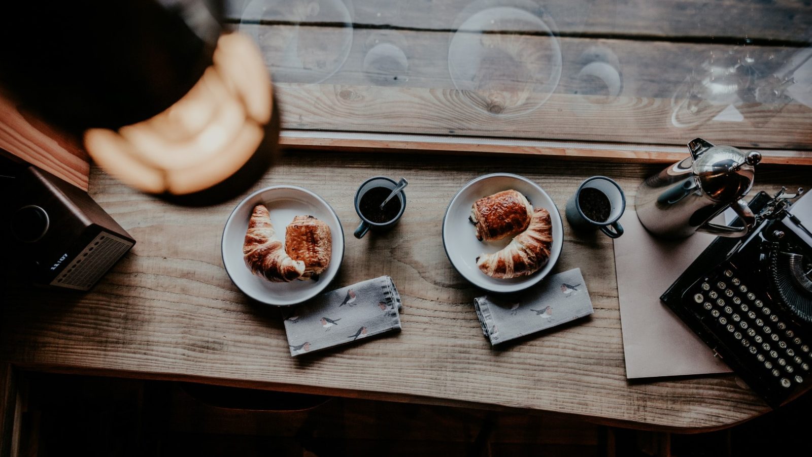 Top view of a wooden table at Little Menherion with two plates of croissants, two cups of coffee, a vintage typewriter, and napkins. Natural light filters through a window, creating a cozy atmosphere.