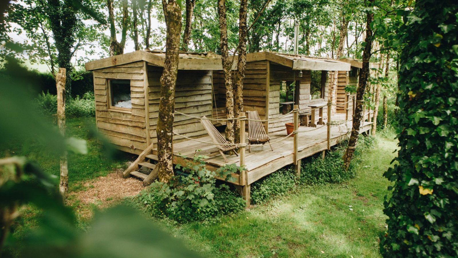 Tucked away in Little Menherion, a rustic wooden treehouse with a deck is surrounded by lush green foliage. Two wooden chairs on the deck invite relaxation in this serene, natural setting, as the treehouse blends harmoniously with the forested environment.