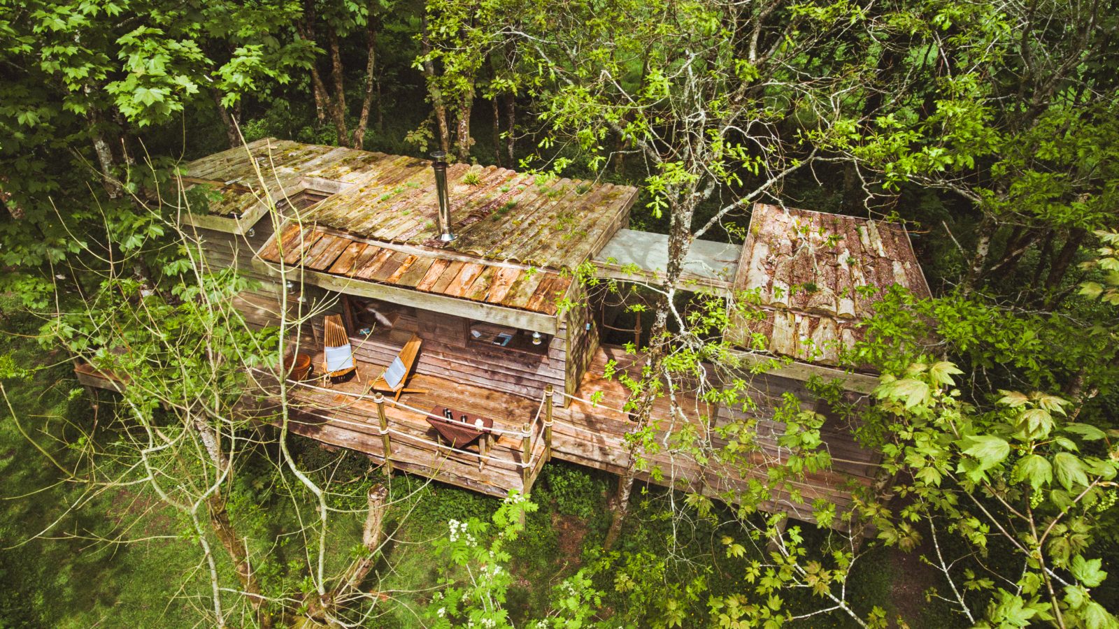 Aerial view of Little Menherion's rustic wooden treehouse nestled among lush green trees. The treehouse features a small deck with two wooden chairs and a bench, creating a tranquil, nature-infused retreat as sunlight filters through the dense foliage.