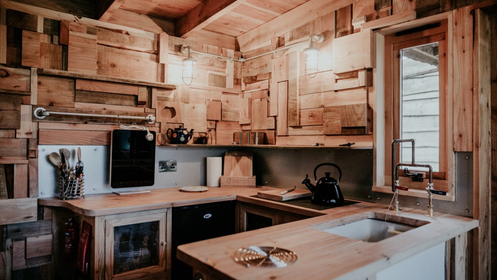 The rustic kitchen at Little Menherion boasts patchwork wood walls, ceiling, and cabinets. It features a countertop with a white sink, a black kettle, and a small cutting board. Soft ambient lighting adds warmth to this cozy enclave.