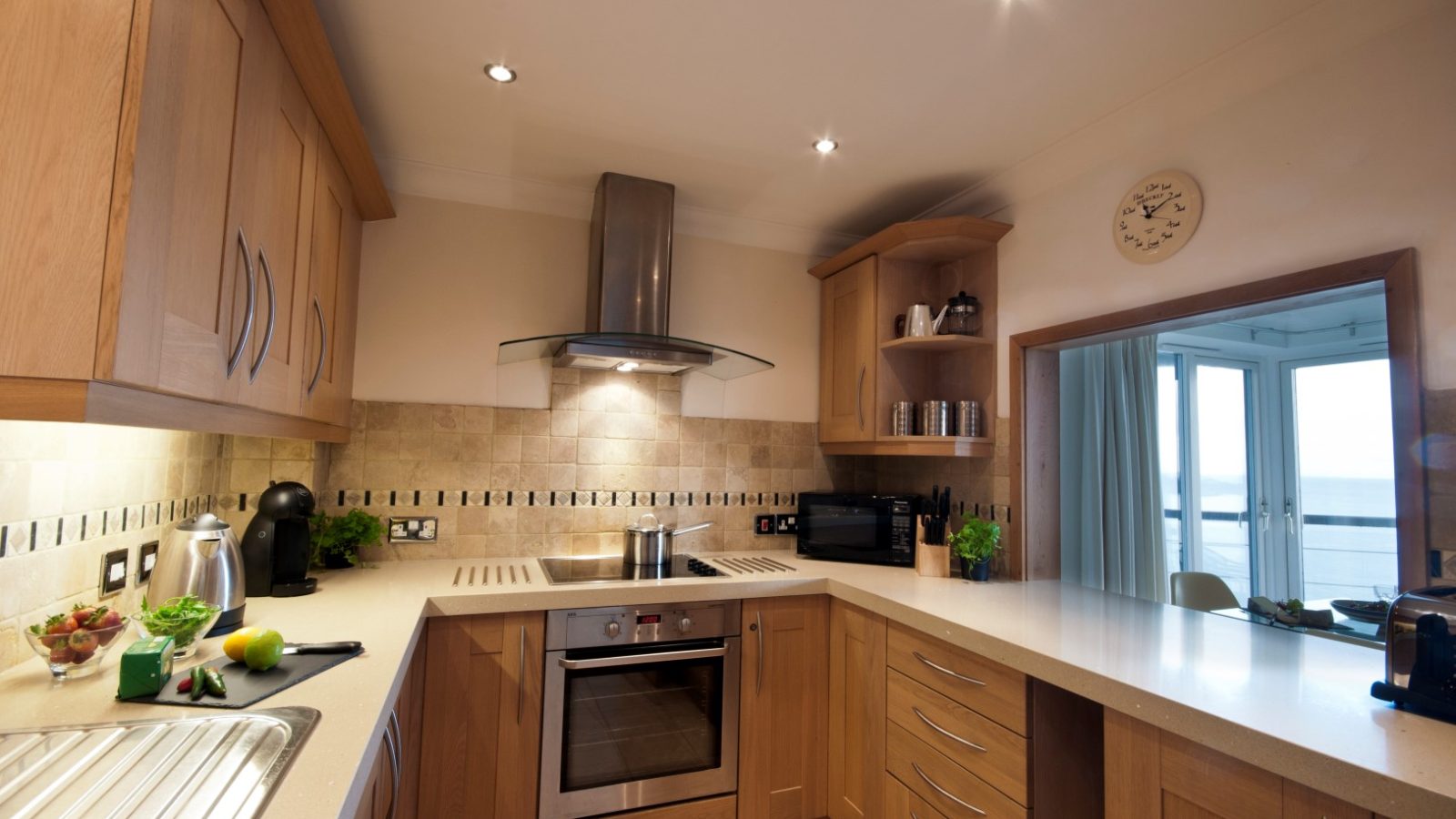 A modern kitchen with wooden cabinets, stainless steel appliances, and a tan tile backsplash exudes a marine charm. It features an electric stove and range hood, while the window offers a sea view. Small potted plants add greenery on the countertop, creating a perfect focal point of tranquility.