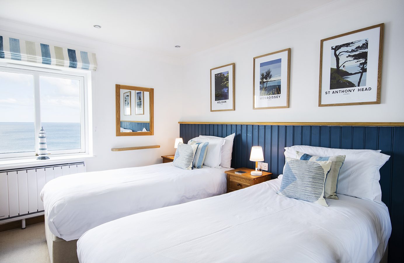 A bright bedroom with two single beds, blue and white striped cushions, and a nautical theme. The window offers a Marine Point ocean view, complemented by wooden furniture and framed coastal pictures. A lamp sits on the nightstand, enhancing the serene ambiance.