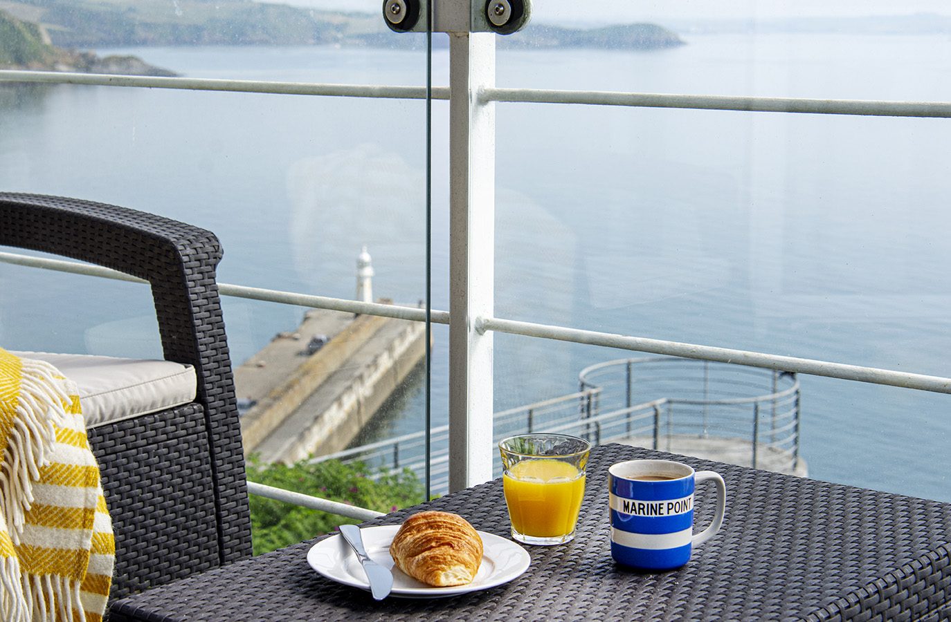 A wicker chair with a yellow and white blanket sits beside a small table on the balcony of Marine Point, overlooking the sea. The table holds a plate with croissants, a glass of orange juice, and a mug labeled 