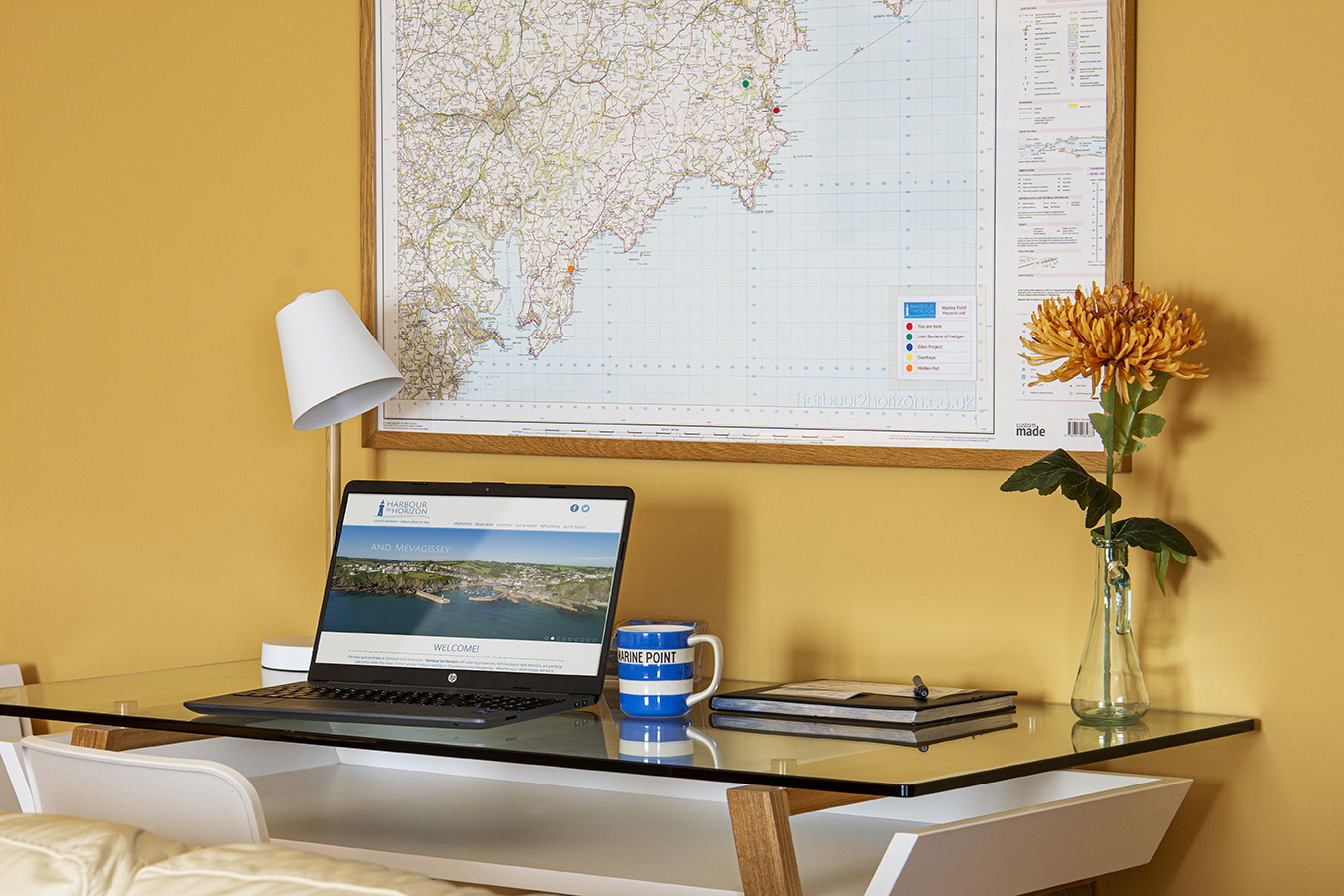 A modern workspace featuring a laptop on a glass desk, with a marine blue mug, notebook, and pen. A large map is mounted on a mustard yellow wall behind the desk, with a white lamp and a single yellow flower in a vase completing this serene Marine Point setup.