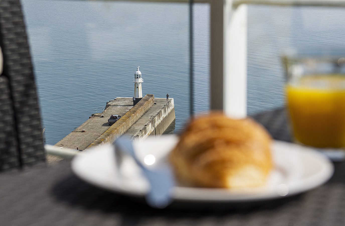 A croissant and glass of orange juice sit on a table in the foreground, with a blurred view of Marine Point's pier and lighthouse extending into the calm sea beyond.