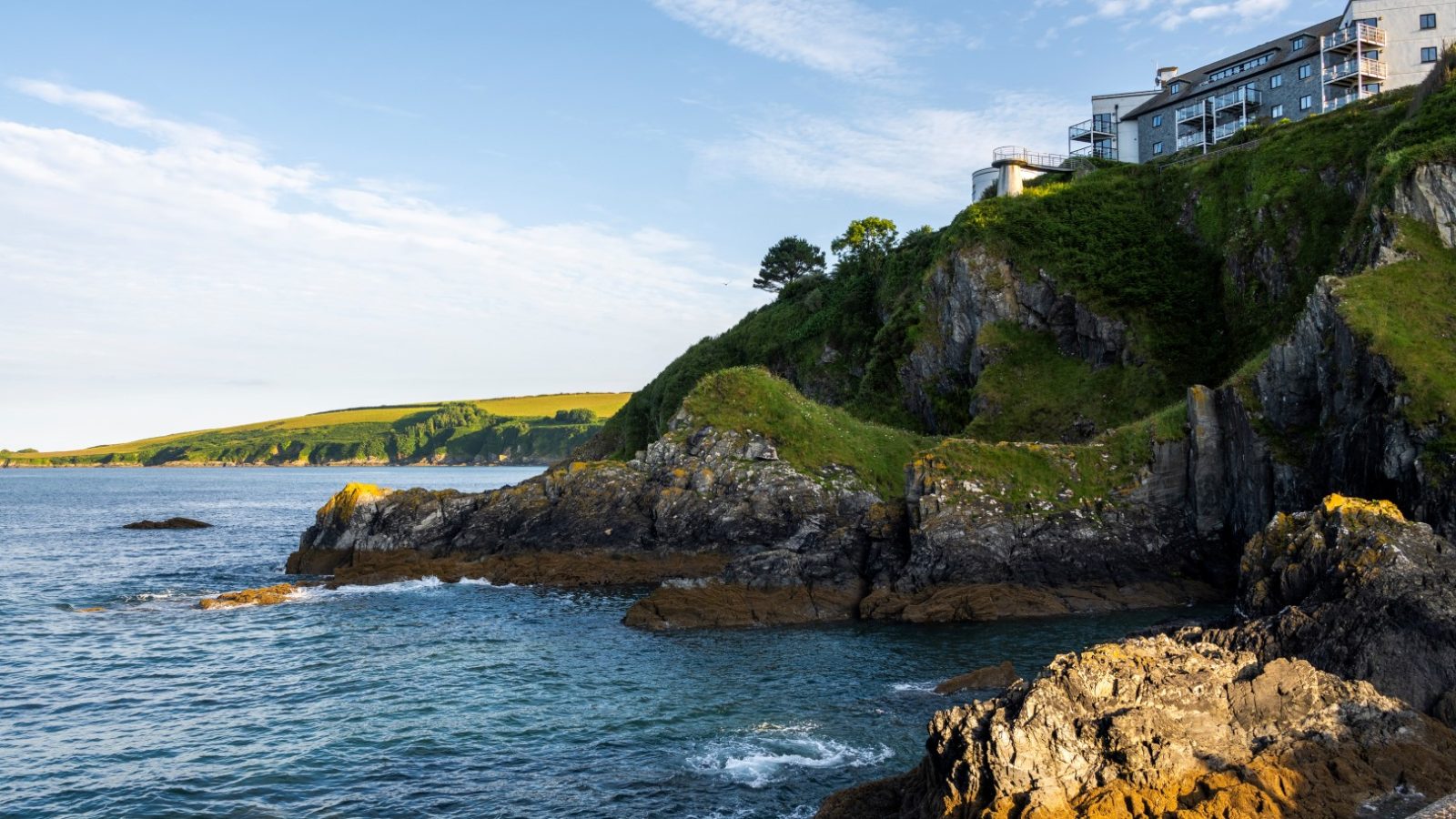 A scenic coastal view shows rocky cliffs, with waves crashing below in a marine spectacle. A few modern buildings point skyward atop the greenery-covered cliffs, overlooking the expansive sea and distant coastline under a clear, blue sky.